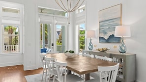 Un coin repas avec une table en bois, des chaises blanches, un saladier rempli de fruits et un tableau représentant une scène de plage.
