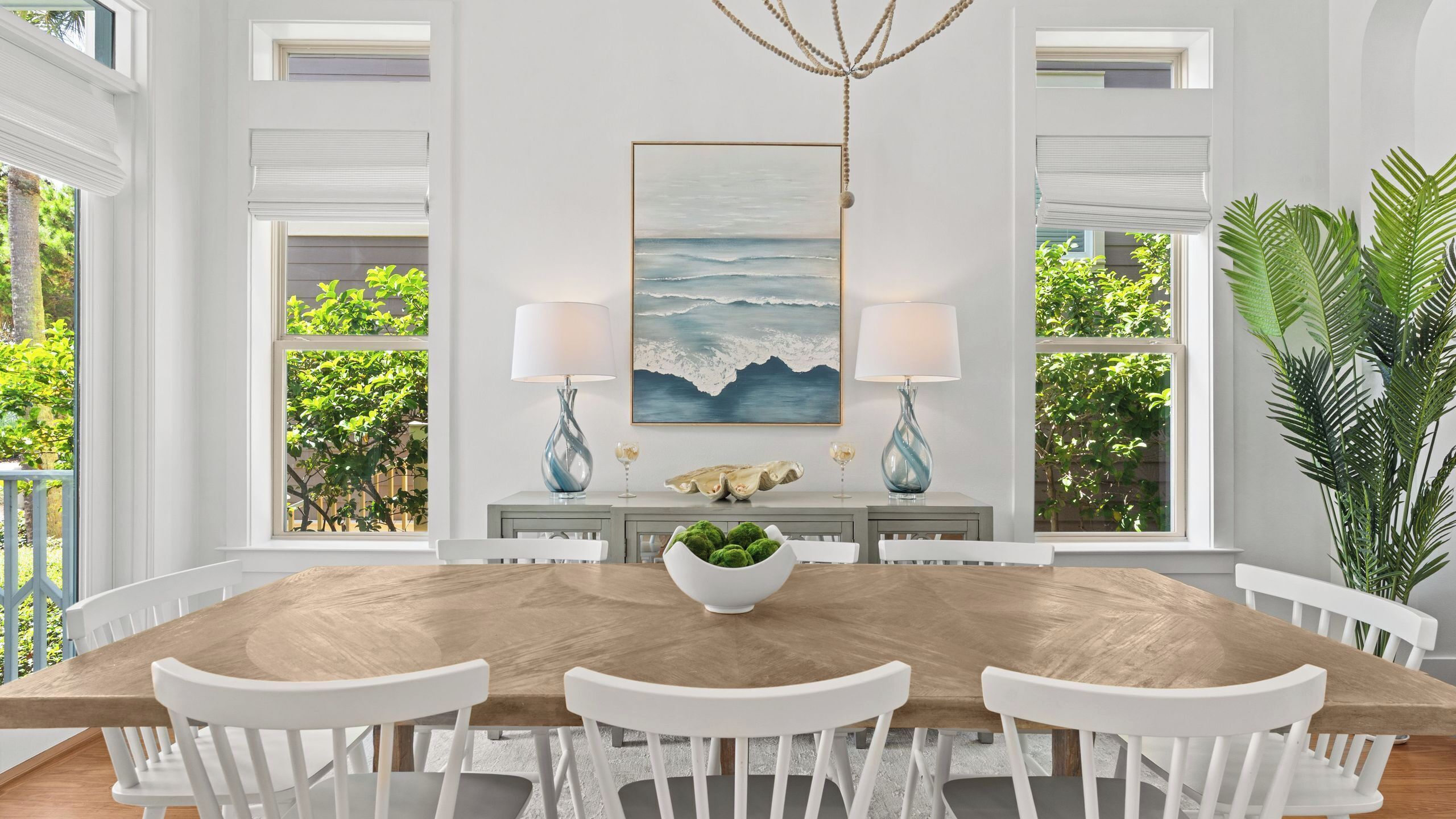 A dining room with a wooden table, white chairs, a central island with a bowl, two lamps, and a large ocean-themed painting on the wall.
