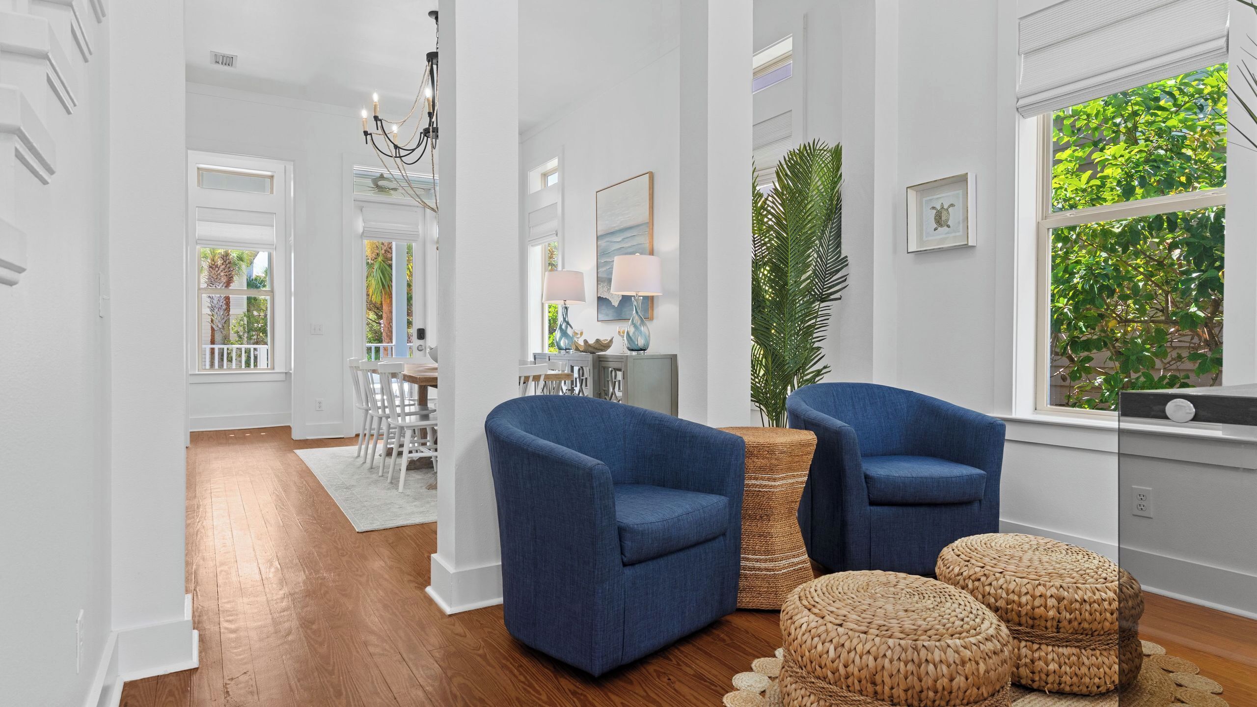 Amplia sala de estar con piso de madera, dos sillones azules, un otoman tejido, un comedor con mesa y sillas blancas, y un ventanal con vegetación en el exterior.
