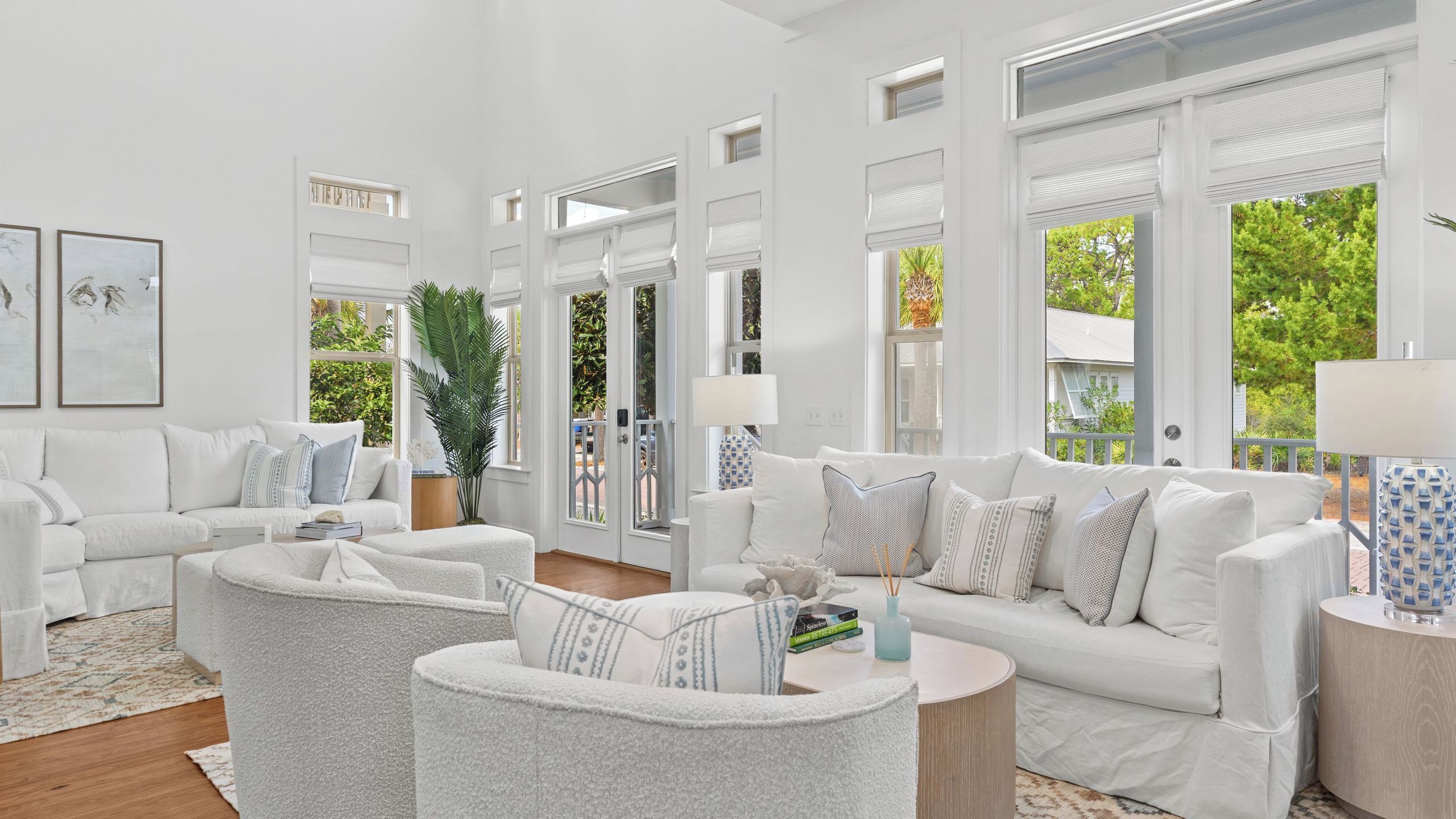 A spacious living room with white furniture, large windows, and a view of greenery outside.