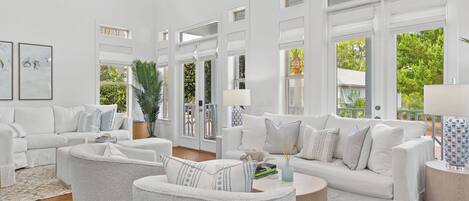 A spacious living room with white furniture, large windows, and a view of greenery outside.