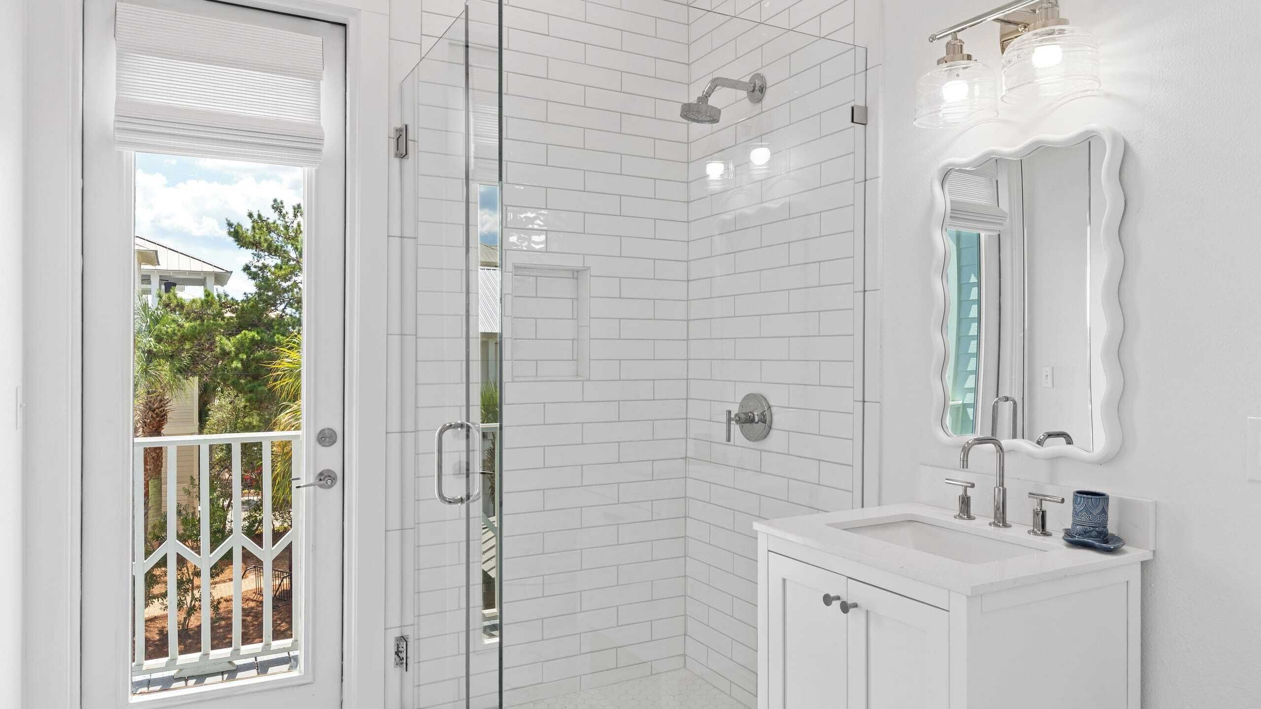 A bathroom with white subway tiles, a glass shower enclosure, a white vanity with a mirror, and a door leading to a balcony with a view of trees and a house.