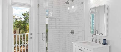 A bathroom with white subway tiles, a glass shower enclosure, a white vanity with a mirror, and a door leading to a balcony with a view of trees and a house.