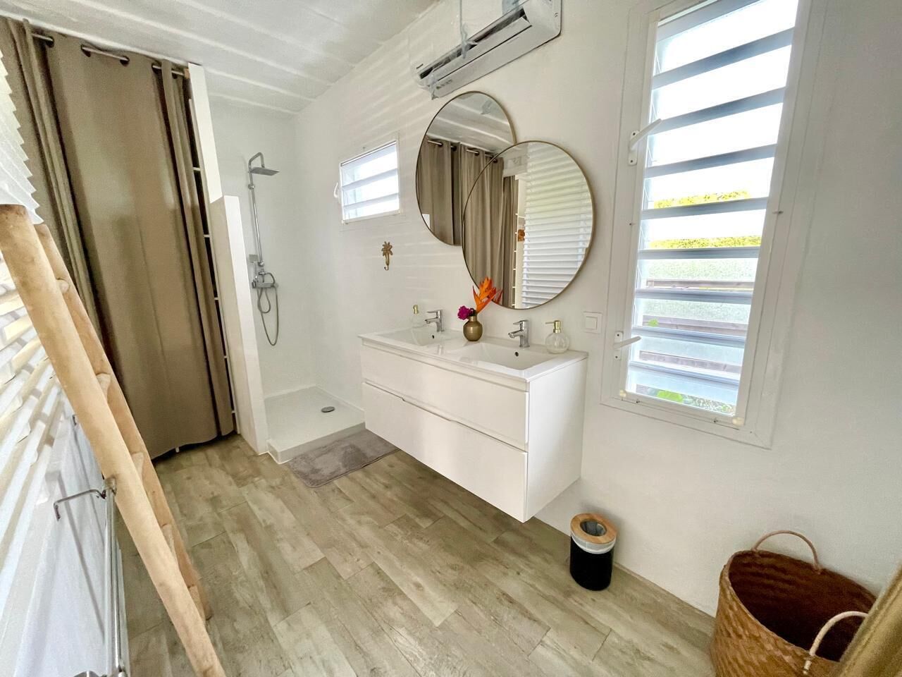 A modern bathroom with a white vanity, two mirrors, a shower, and a window with blinds.