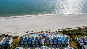 Vue aérienne d’un hôtel en bord de mer, avec plusieurs bâtiments aux toits bleus, une plage de sable et une vue dégagée sur l’océan.