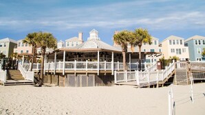 Un belvédère au bord de la plage, doté d’un espace salon couvert, entouré de palmiers et bordé d’une rangée de maisons de plage.