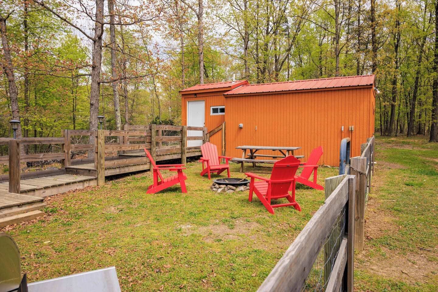 A wooden cabin with a red roof, a picnic table, and red Adirondack chairs in a wooded area.