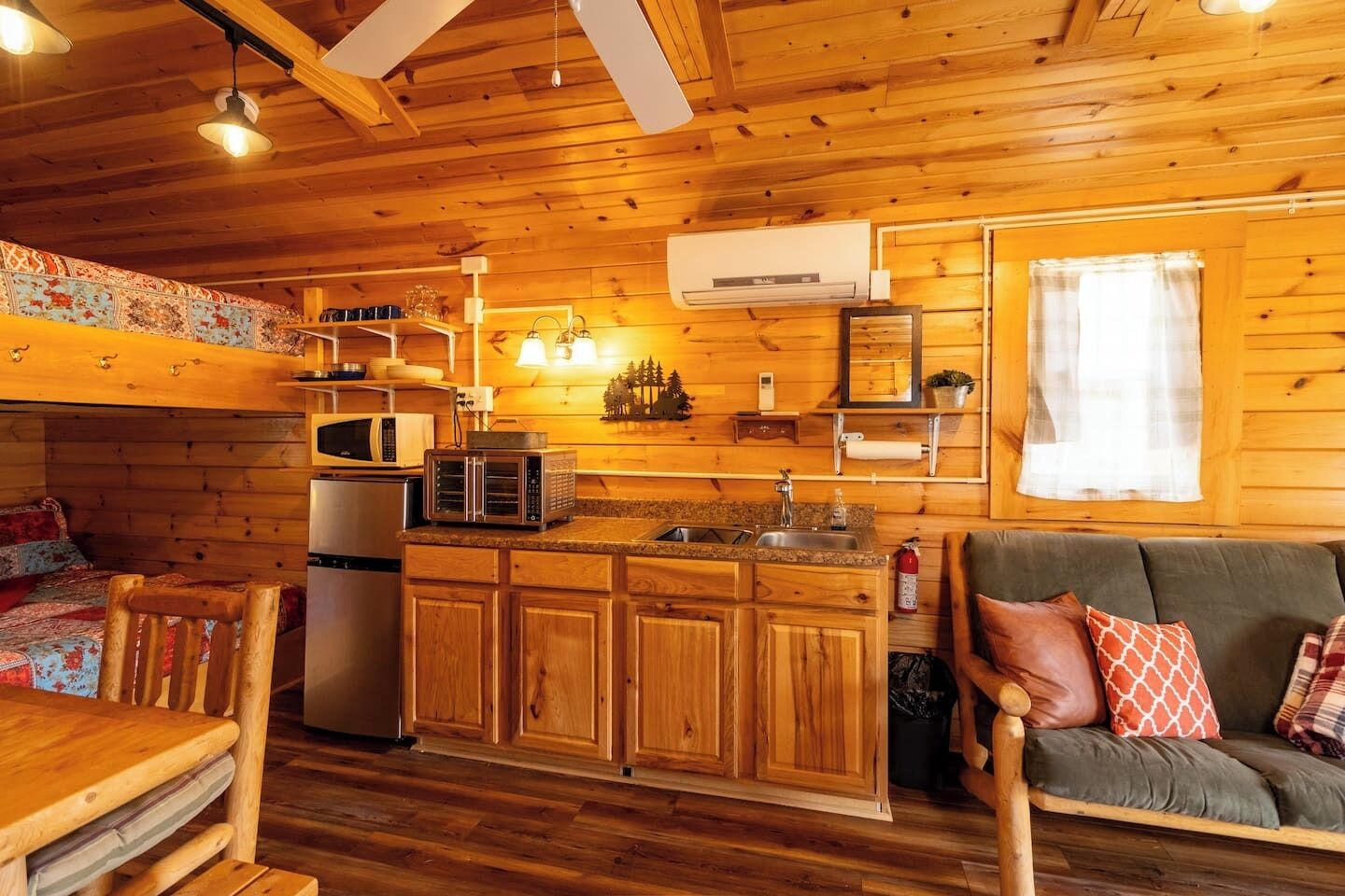 A cozy cabin-style interior with wooden walls and floors, a kitchenette, a dining area with a table and chairs, and a sofa with cushions.