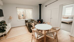 A cozy living room with a round table and chairs, a sofa, a white cabinet, a large window with a view of trees, and a black wood-burning stove. - Salt Cottage - charming two–bedroom retreat (Port Fairy)