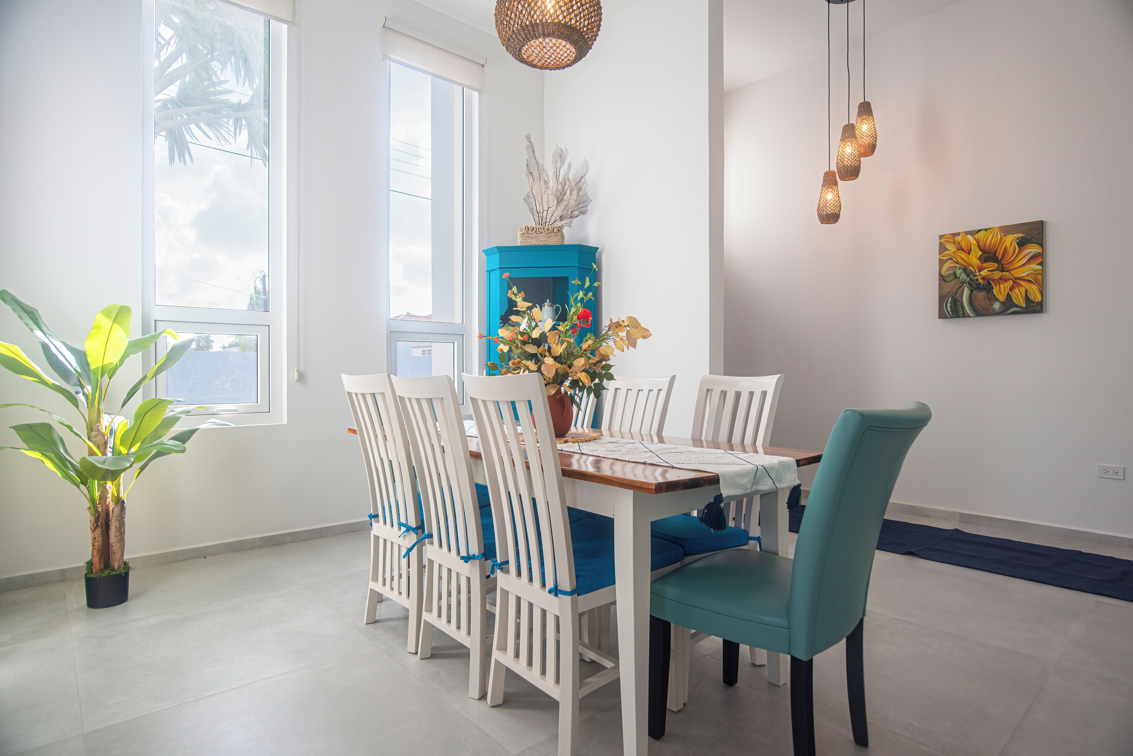 Un coin repas avec une table en bois, des chaises blanches et une chaise de couleur turquoise. On y trouve un meuble bleu sur lequel est posé un objet décoratif, une plante en pot et un tableau accroché au mur.