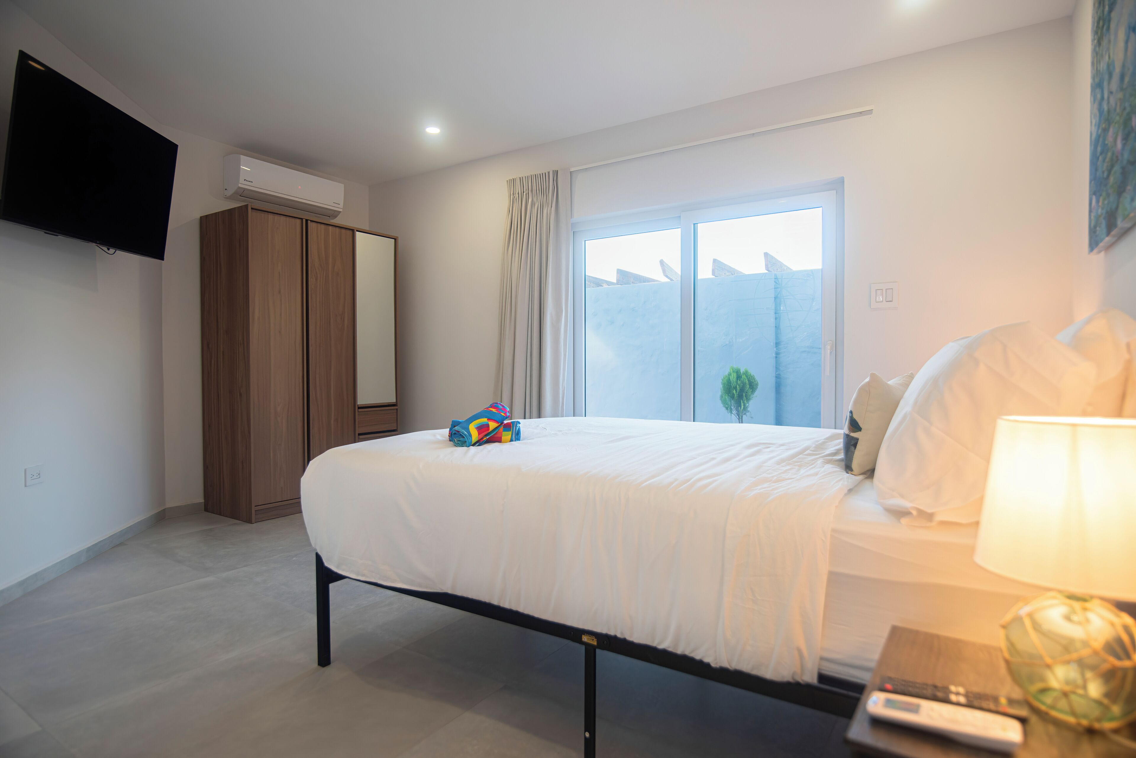 Een slaapkamer met een bed, een aan de muur bevestigde televisie, een airconditioner, een raam met gordijnen en een nachtkastje met een lamp.