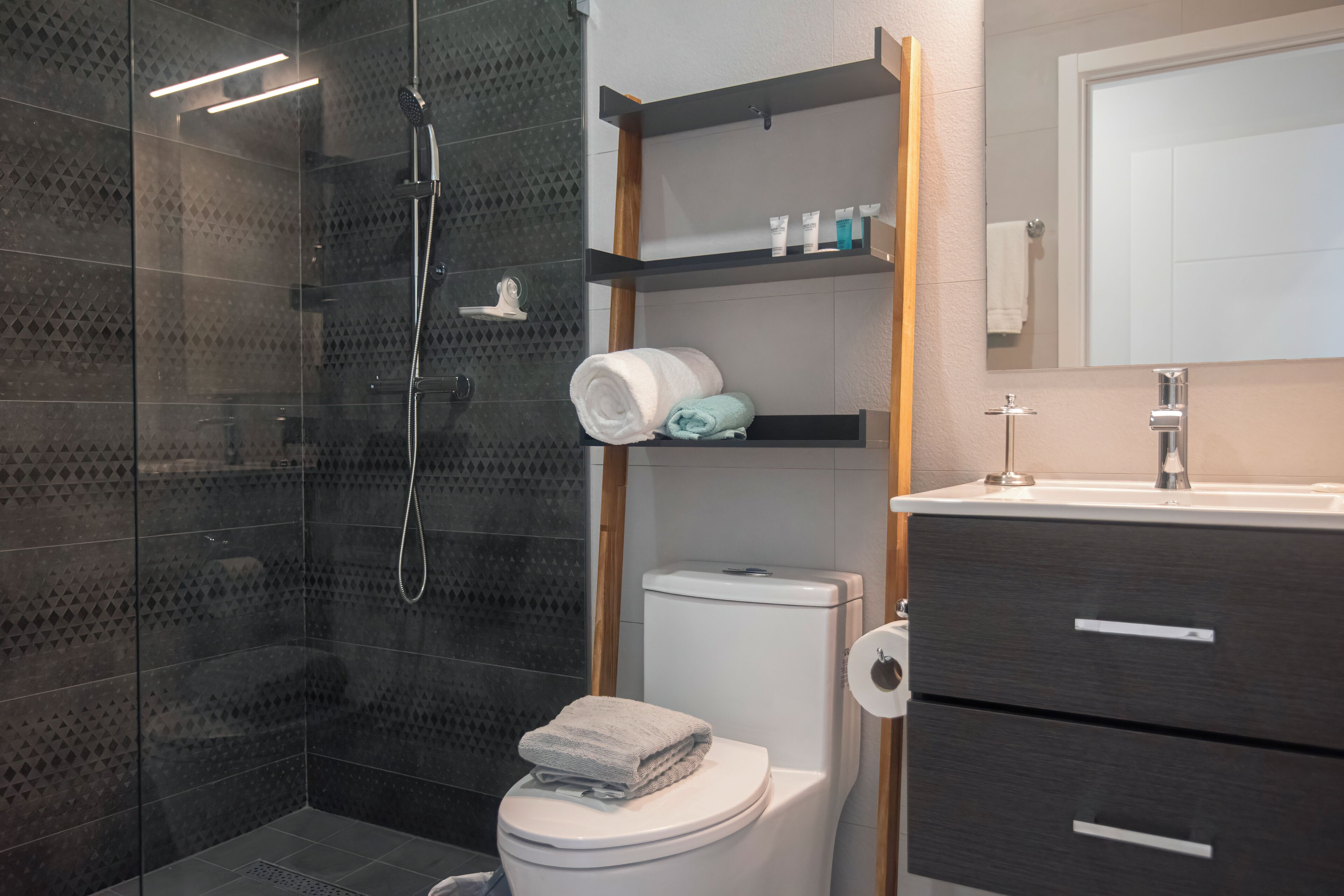 A modern bathroom with a glass-enclosed shower, a toilet with a towel on top, and a vanity with a mirror and shelves.