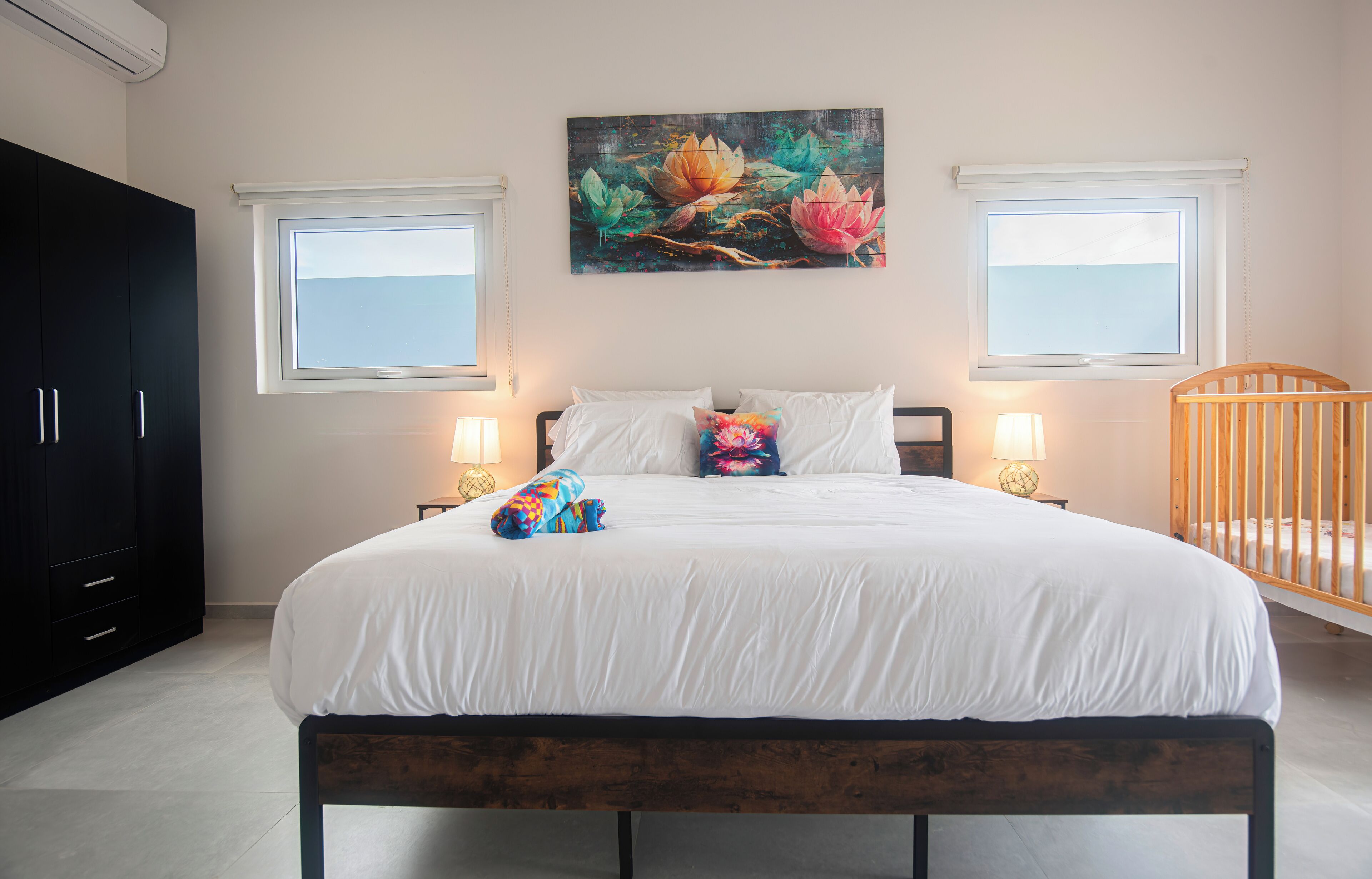Un dormitorio con una cama grande, dos lámparas de noche, una cuna y un cuadro de flores en la pared.