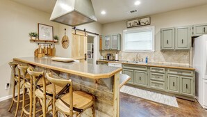 Una cocina con una isla central, taburetes de bar, armarios verde claro y un ventanal con persianas.