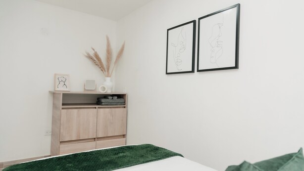 A minimalist bedroom with a bed, a green throw, a wooden wardrobe, and two framed artworks on the wall.