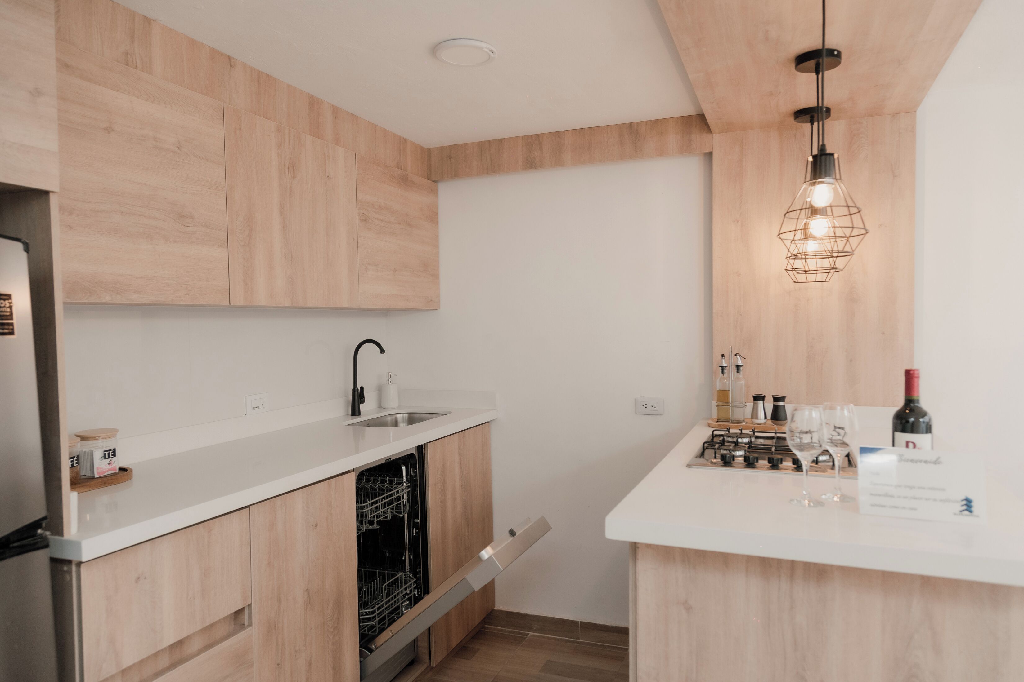 Una cocina moderna con armarios de madera, encimera blanca, lavavajillas, fregadero y una botella de vino.