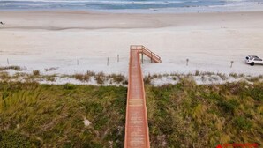 A beach with a wooden boardwalk leading to the water, a white vehicle parked on the sand, and a clear sky.