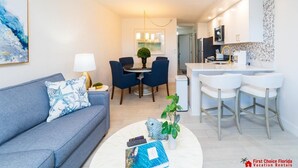 A modern living room with a grey sofa, a coffee table, and a dining area with blue chairs. There is a kitchen area with white cabinets and a white countertop.