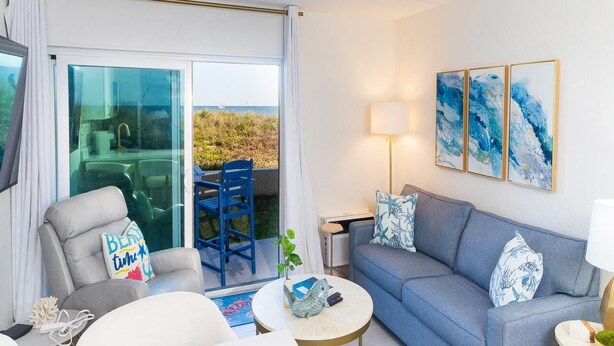 A modern living room with a sofa, armchair, and coffee table. There is a sliding glass door leading to a balcony with a view of the ocean.