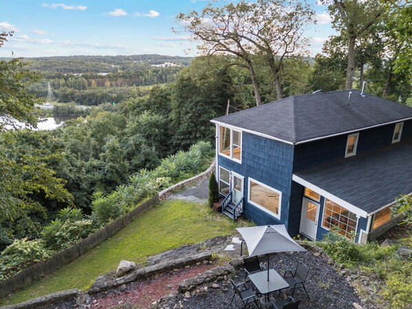 A two-story house with a dark blue exterior, a large window on the second floor, and a view of a lake and forest in the background. - Casa Mattia 4BR Luxury Mountain Home (Suffern)