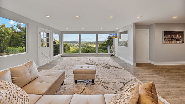 A spacious living room with a large window, a beige sofa, an ottoman, and a marble-patterned rug. - Casa Mattia 4BR Luxury Mountain Home (Suffern)
