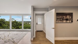 A hallway with a view of greenery and a cityscape through large windows, a wooden floor, and a door leading to another room. - Casa Mattia 4BR Luxury Mountain Home (Suffern)