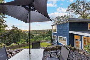 A patio with a table and chairs, a large umbrella, and a view of a blue house with a deck and a stone wall. - Casa Mattia 4BR Luxury Mountain Home (Suffern)