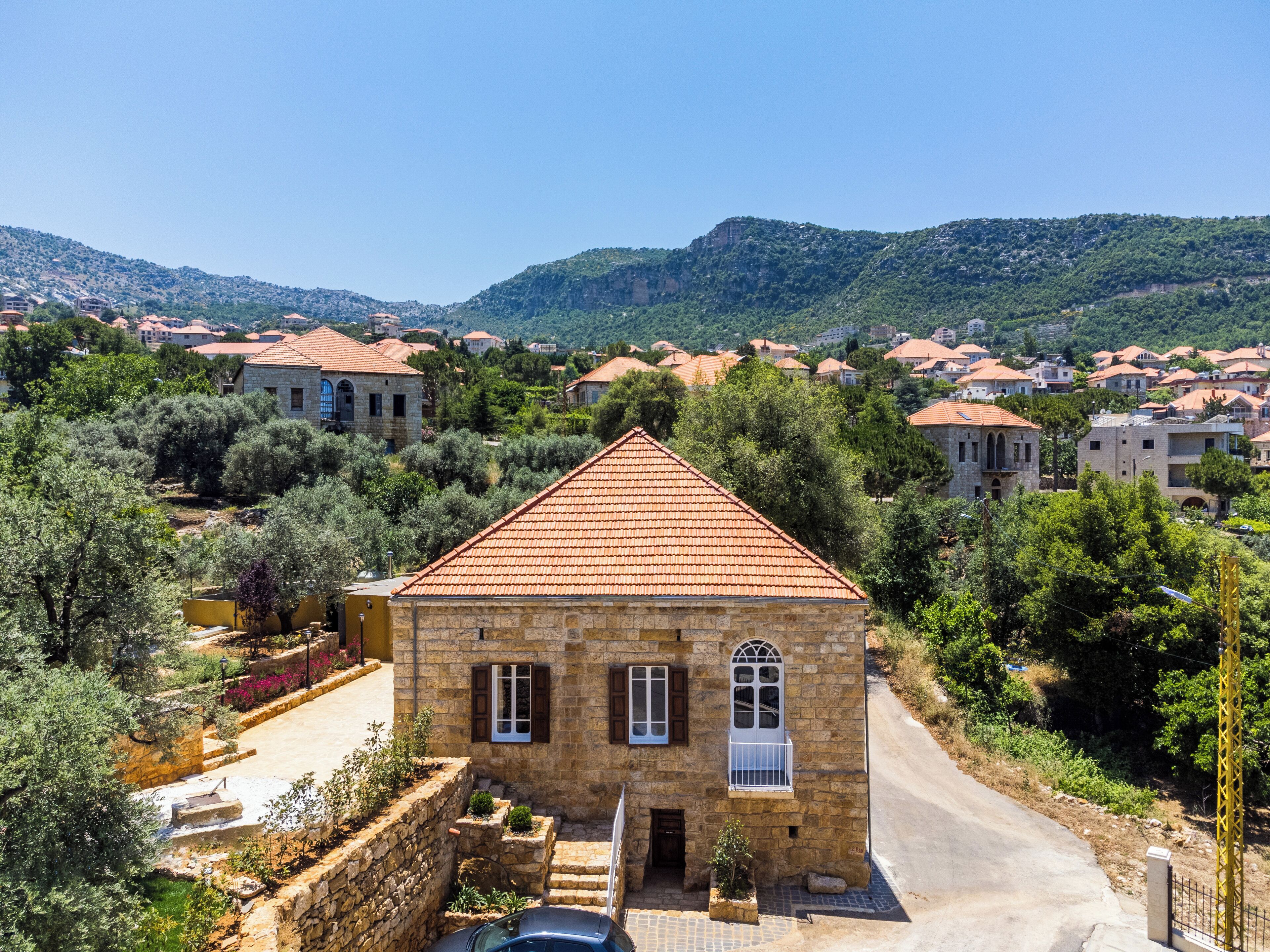 Um edifício de pedra com telhado em telhas, rodeado por oliveiras e com montanhas ao fundo.