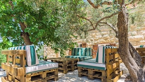 Espace extérieur aménagé avec des meubles en bois et des coussins, adossé à un mur de pierre et abrité par des arbres.