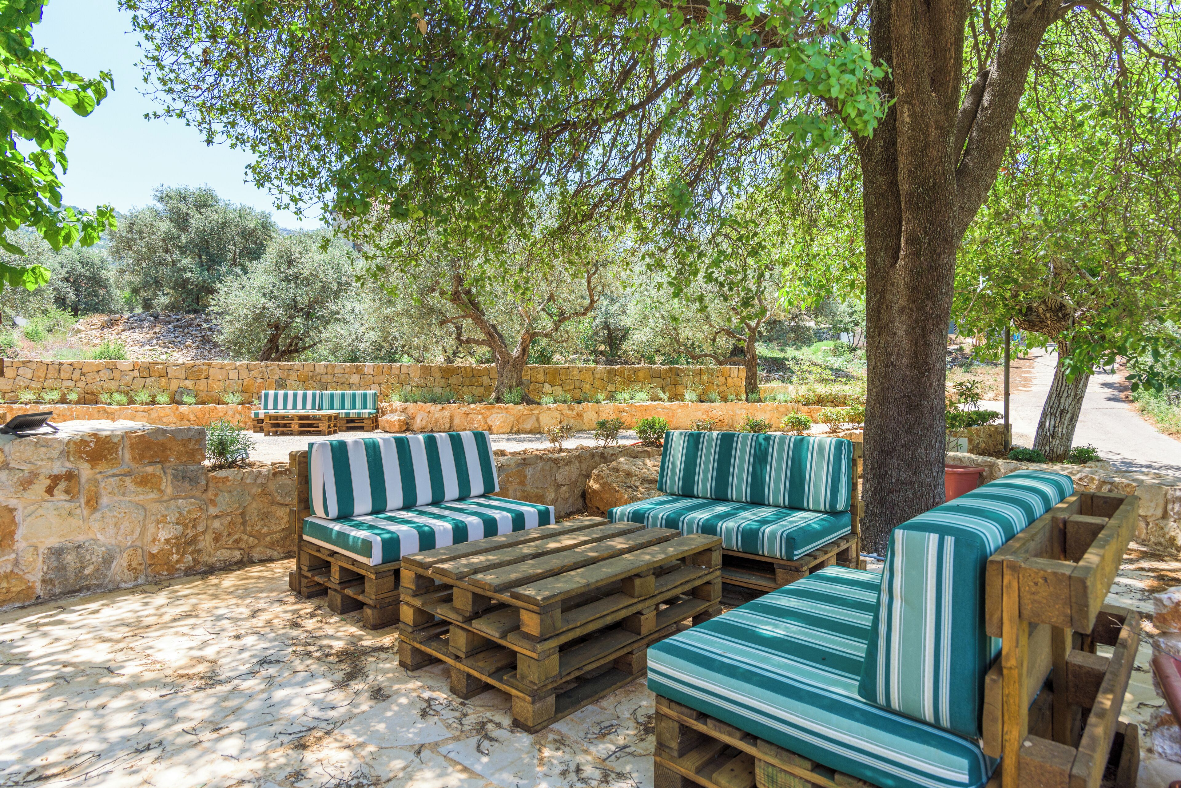 An outdoor seating area with wooden furniture and striped cushions, surrounded by stone walls and trees.