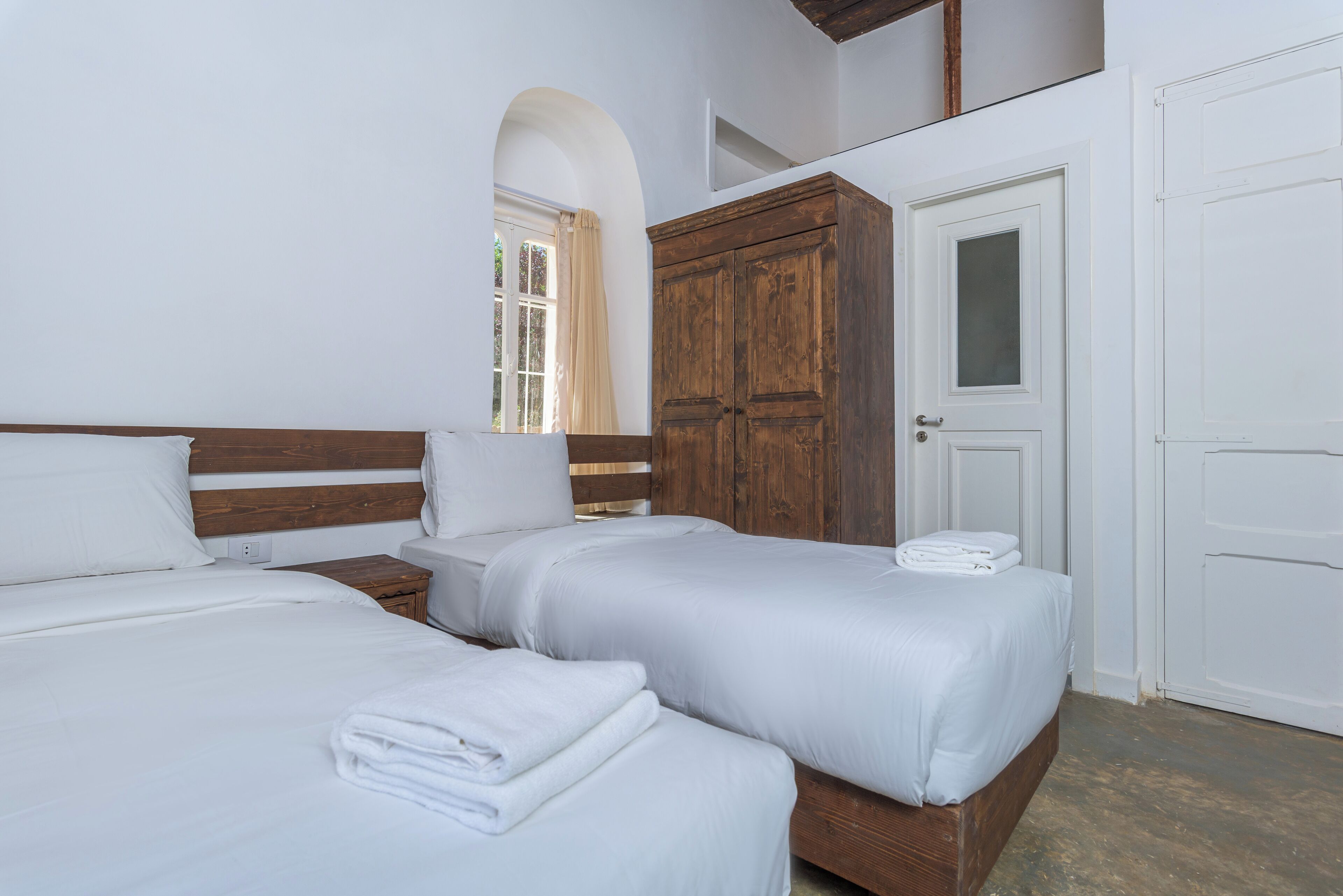 A hotel room with two beds, a wooden wardrobe, a white door, and an arched window.