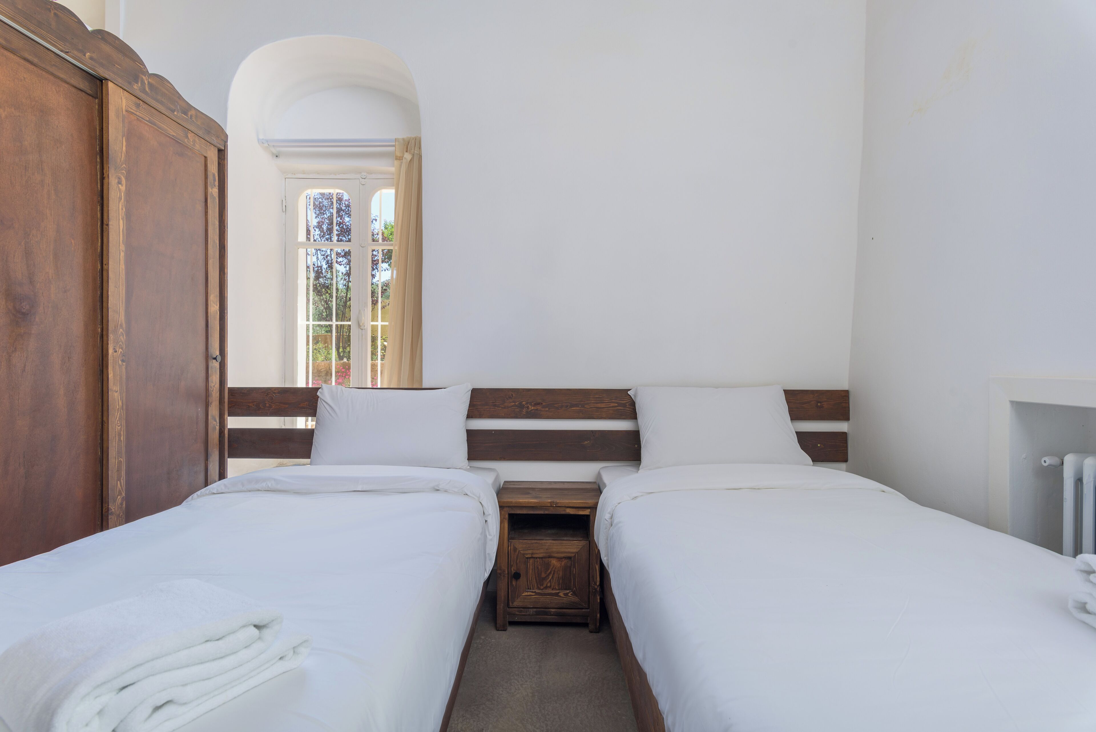 Una habitación con dos camas individuales, cabeceros de madera, ropa de cama blanca, una mesita de noche de madera y una ventana con cortinas.