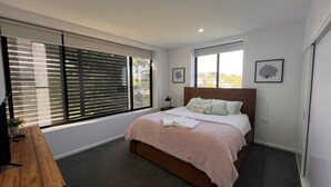 Une chambre moderne avec un grand lit, un bureau en bois et une vue sur la ville grâce à de grandes fenêtres du sol au plafond.