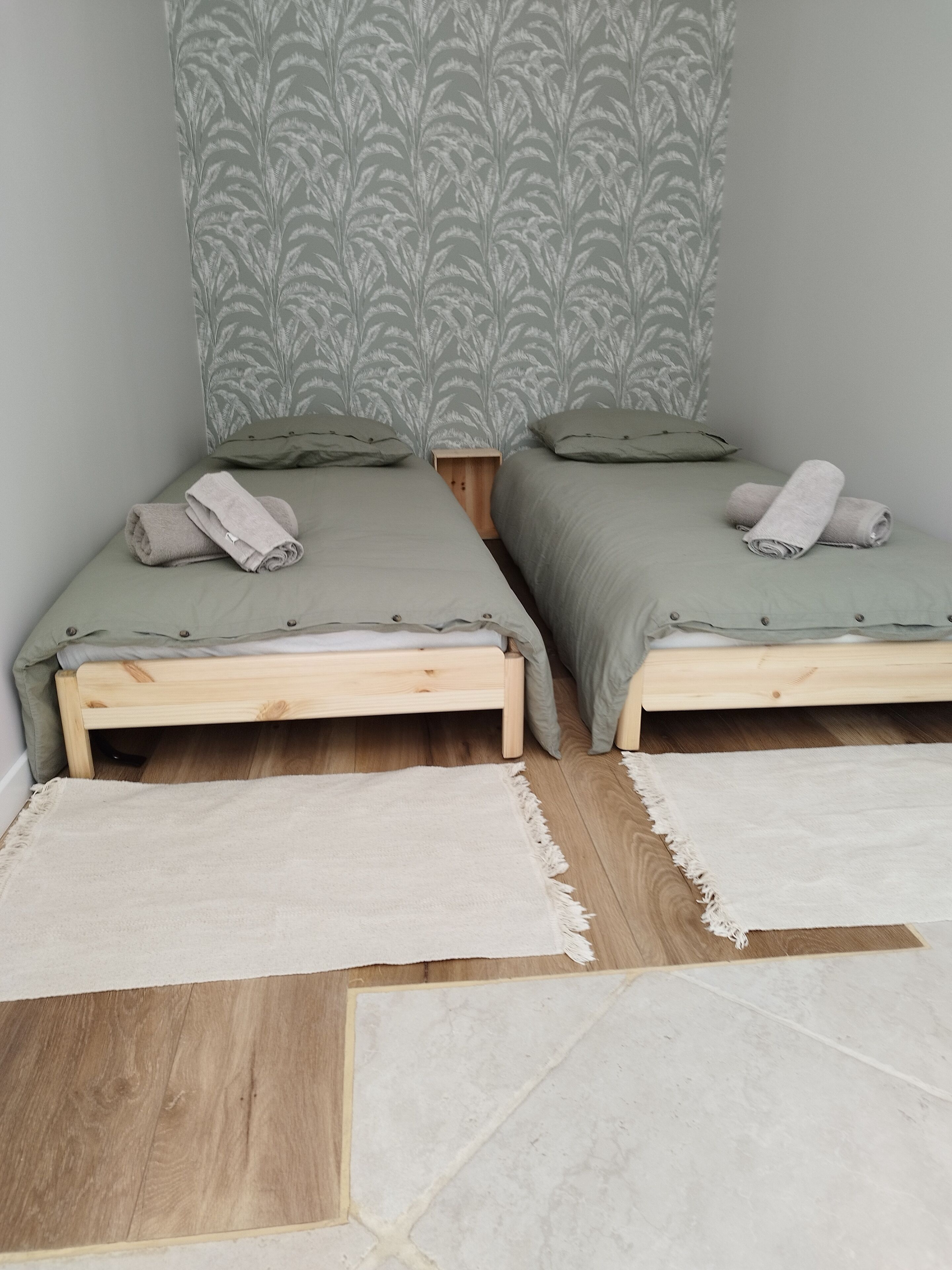 Une chambre avec deux lits en bois, chacun recouvert d’une couverture verte et agrémenté d’oreillers, et un papier peint à motifs sur le mur.