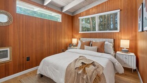 A bedroom with wooden paneling, a large bed with white bedding, two bedside tables with lamps, and a window with blinds. - Mid-Century Hideaway, Near Forest Park (Portland)