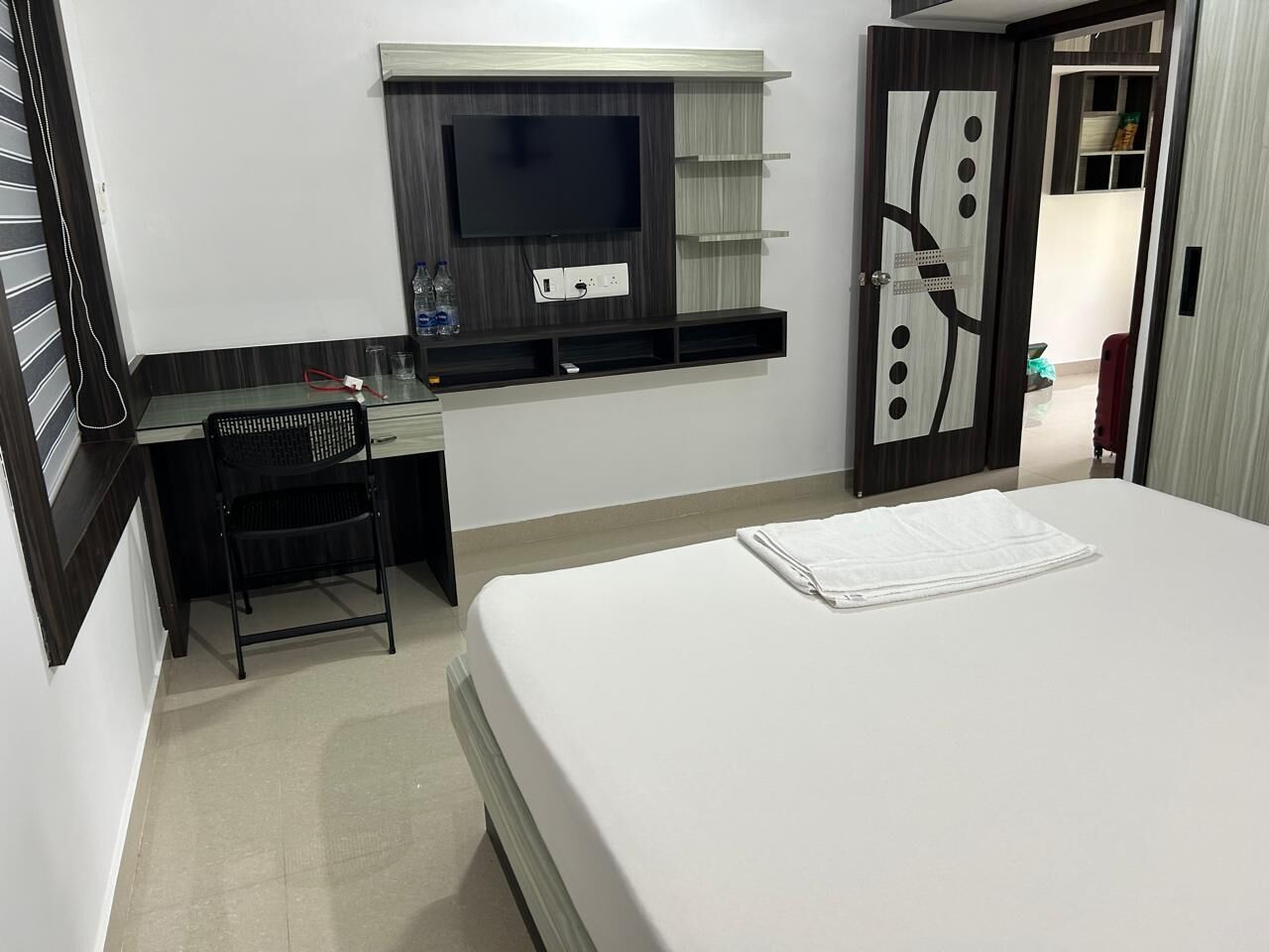 A hotel room with a bed, a desk with a chair, a TV mounted on the wall, and a wardrobe with a mirror.