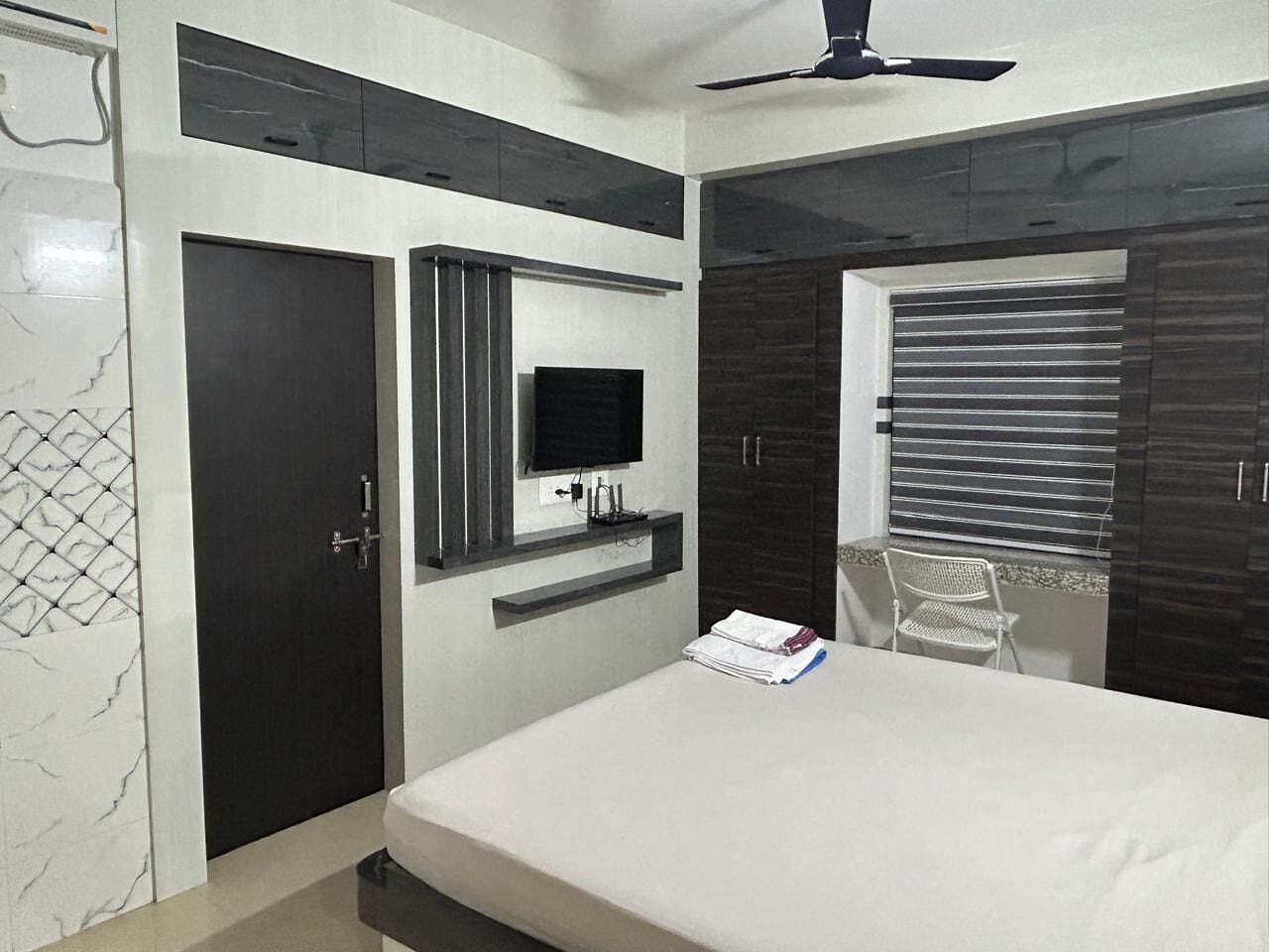 A modern bedroom with a bed, a TV mounted on the wall, a wardrobe, and a window with blinds.