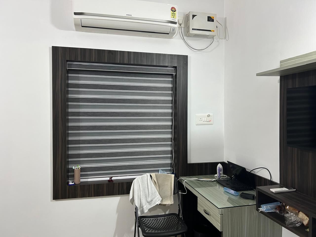 Et moderne værelse med et stort vindue med vandrette persienner, et skrivebord med en bærbar computer, en stol og en aircondition-enhed monteret på væggen.