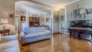 A spacious living room with a grand piano, a sofa, and a dining area with wooden cabinets and framed artwork. - Roman Holiday Republic (Roma)
