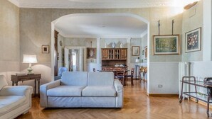 A spacious living room with a light-colored sofa, wooden flooring, and a kitchen area in the background. - Roman Holiday Republic (Roma)