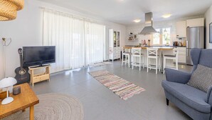 Une cuisine moderne avec des meubles blancs, un coin repas avec des chaises blanches, un canapé gris et un téléviseur posé sur un meuble en bois.