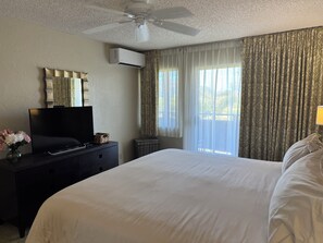 Una habitación de hotel con cama, televisión en una cómoda, aire acondicionado, ventilador de techo y ventana con cortinas.