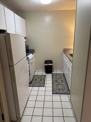Cocina compacta con gabinetes blancos, un cubo de basura negro y alfombras estampadas sobre el suelo de baldosas.
