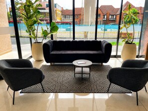 A modern lounge area with a black sofa, two armchairs, a patterned rug, and large windows offering a view of a pool and buildings.