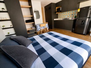 A modern bedroom with a checkered bedspread, a desk with a chair, a kitchen area with a refrigerator, and built-in shelving.