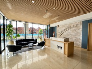 Een modern ingerichte receptie met een houten plafond, een receptiebalie, een zwarte bank en een groot raam met uitzicht op de omgeving.