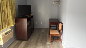 Une petite pièce avec un téléviseur posé sur un meuble en bois, une chaise, une petite table et une étagère fixée au mur.