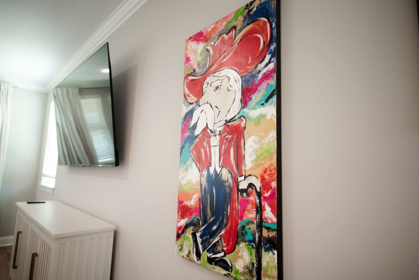 Una habitación con un cuadro grande en la pared, un armario blanco y un televisor de pantalla plana sobre un ventanal con cortinas.