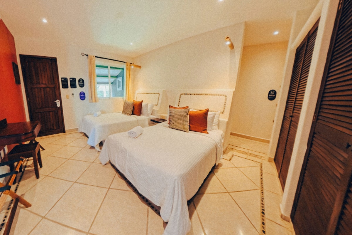 Een hotelkamer met twee bedden, een bureau en een stoel. Er is een houten deur en een raam met gordijnen.