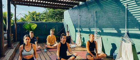 Un grupo de personas sentadas sobre colchonetas de yoga en un espacio al aire libre, con una terraza de madera y una pérgola encima.